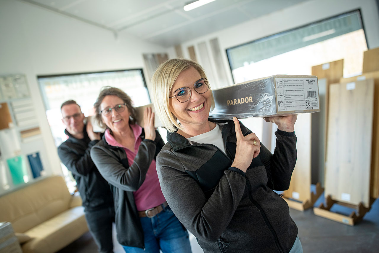 Motiviertes Team bei bodenverkauf.live Gut gelaunte Mitarbeitende tragen ein Paket mit Bodendielen auf den Schultern